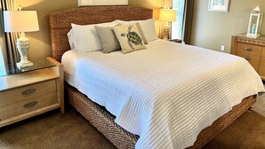 A neatly made bed with a white comforter and several pillows, including a turtle-themed one. There are two bedside tables with lamps, a wooden nightstand, and a small cabinet with drawers. - Beautiful Owner Managed Condo at the Crescent - Best Complex in Destin (Miramar Beach)