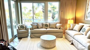 A living room with a beige sectional sofa, a round coffee table, and a view of a balcony with palm trees. - Beautiful Owner Managed Condo at the Crescent - Best Complex in Destin (Miramar Beach)