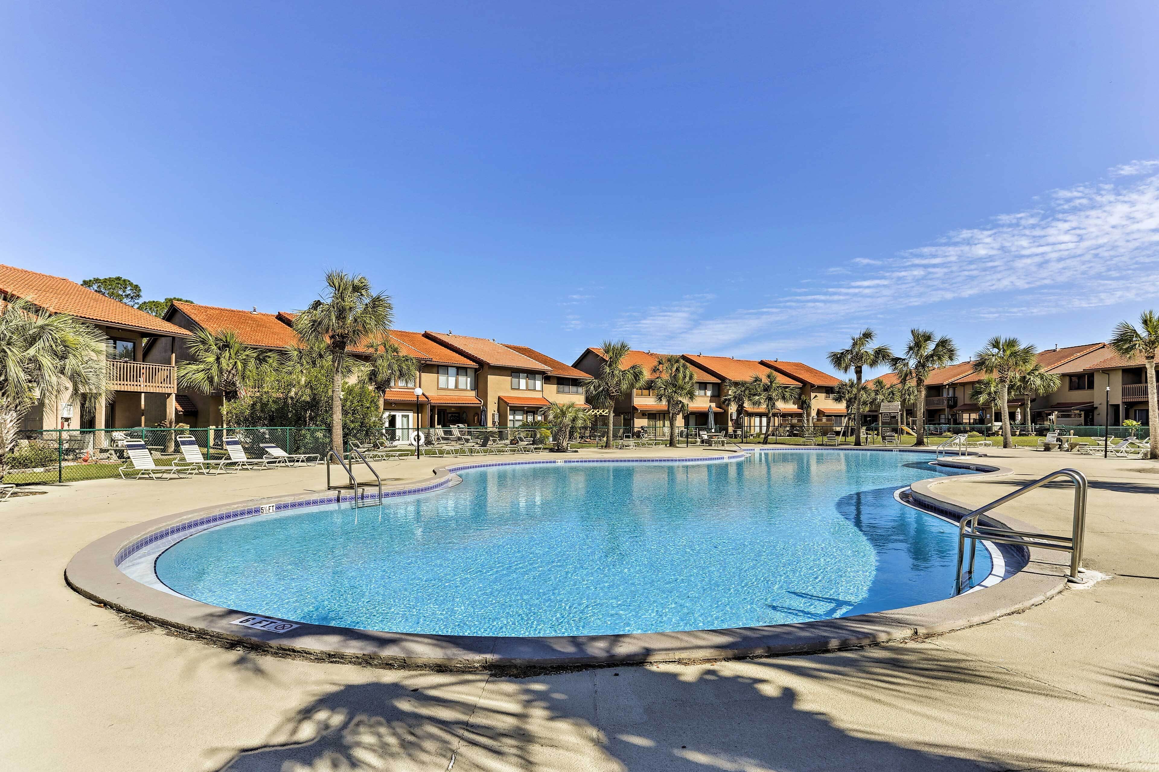 A large, curved swimming pool surrounded by palm trees and buildings with red-tiled roofs.