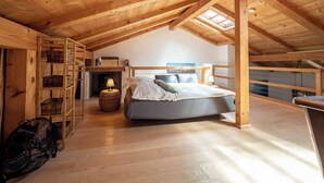 A spacious attic bedroom with a high ceiling, wooden beams, a skylight, a bed, a fireplace, and a fan. - Maisonette apartment for 4 people, 95 sqm with terrace (Obing)