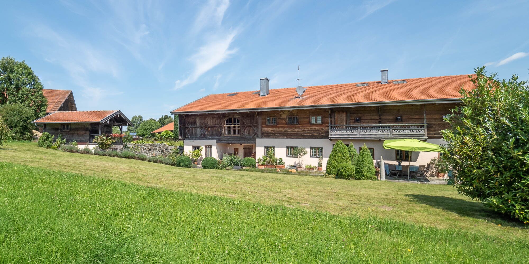 A large, two-story wooden house with a red-tiled roof, surrounded by a well-maintained lawn and a variety of shrubs and trees.