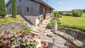 A two-story wooden house with a stone staircase leading to a wooden deck. - Maisonette apartment for 4 people, 95 sqm with terrace (Obing)