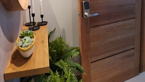 En dörr av trä med ett modernt handtag, en liten hylla med ljus och en växt, och en väggmonterad lampa.