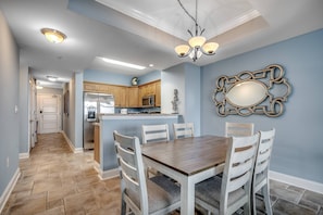 A dining area with a wooden table and chairs, a chandelier, and a kitchen in the background.