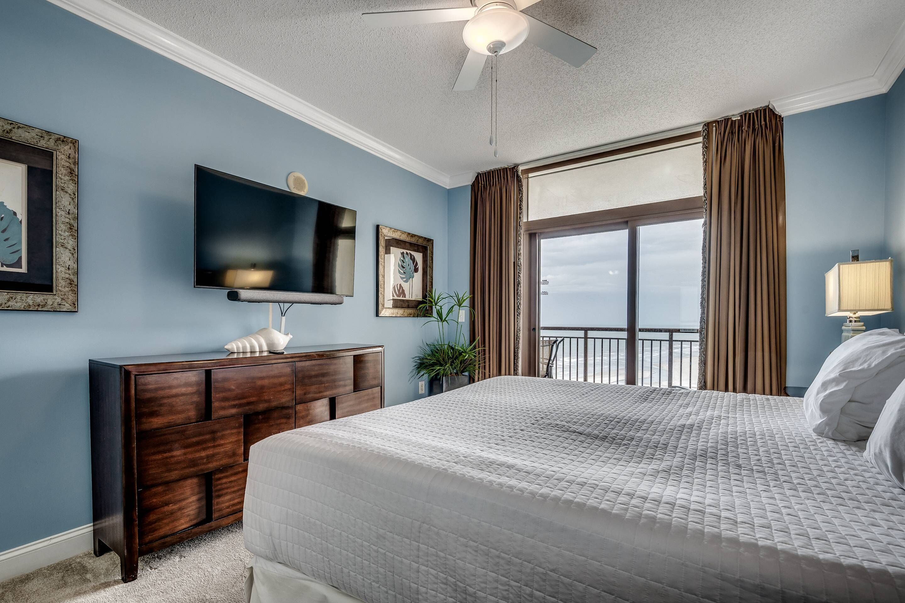 A bedroom with a large bed, a dresser, a flat-screen TV, and a balcony with curtains.