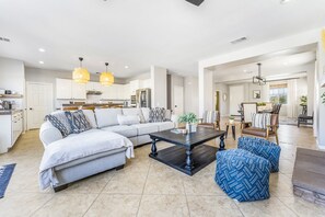 Ein geräumiges Wohnzimmer mit einer Couch, einem Couchtisch und Hocker. Im Hintergrund befindet sich eine Küche mit weißen Schränken und ein Essbereich mit Stühlen.