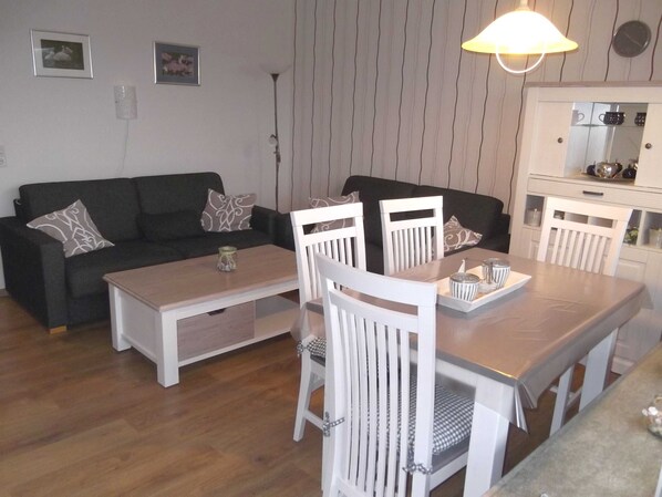 Un salon avec un canapé foncé, des chaises blanches, une table basse en bois et un coin cuisine doté de placards et d’un luminaire suspendu.