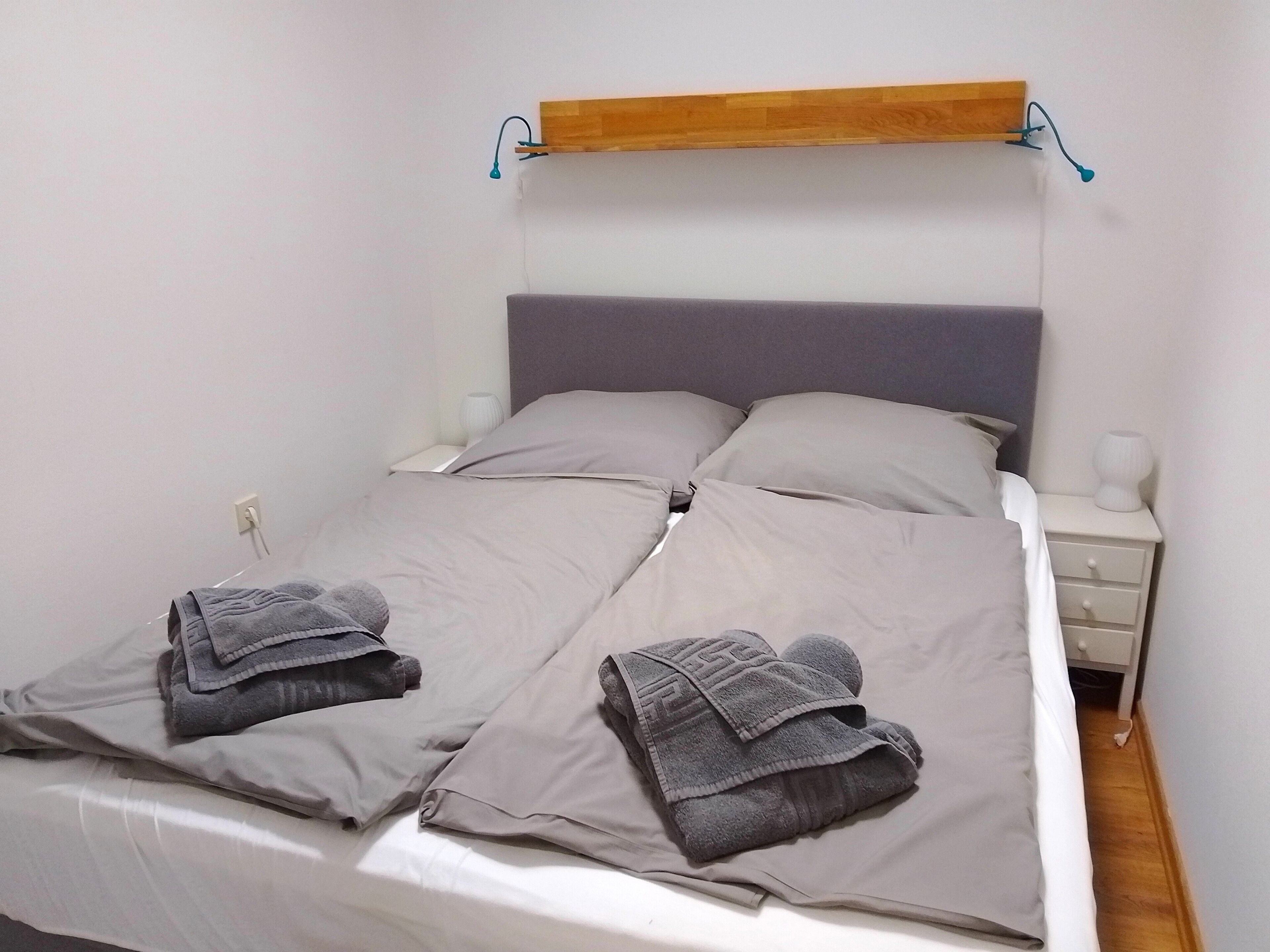 Ein ordentlich bezogenes Bett mit zwei gefalteten Handtüchern, einer Holzablage darüber und einem Nachttisch mit einer Lampe.