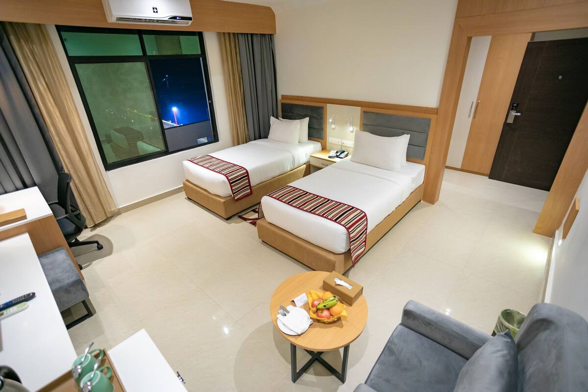 Quarto de hotel com duas camas, uma escrivaninha, uma cadeira, uma mesinha de centro com uma fruteira e uma janela com cortinas.