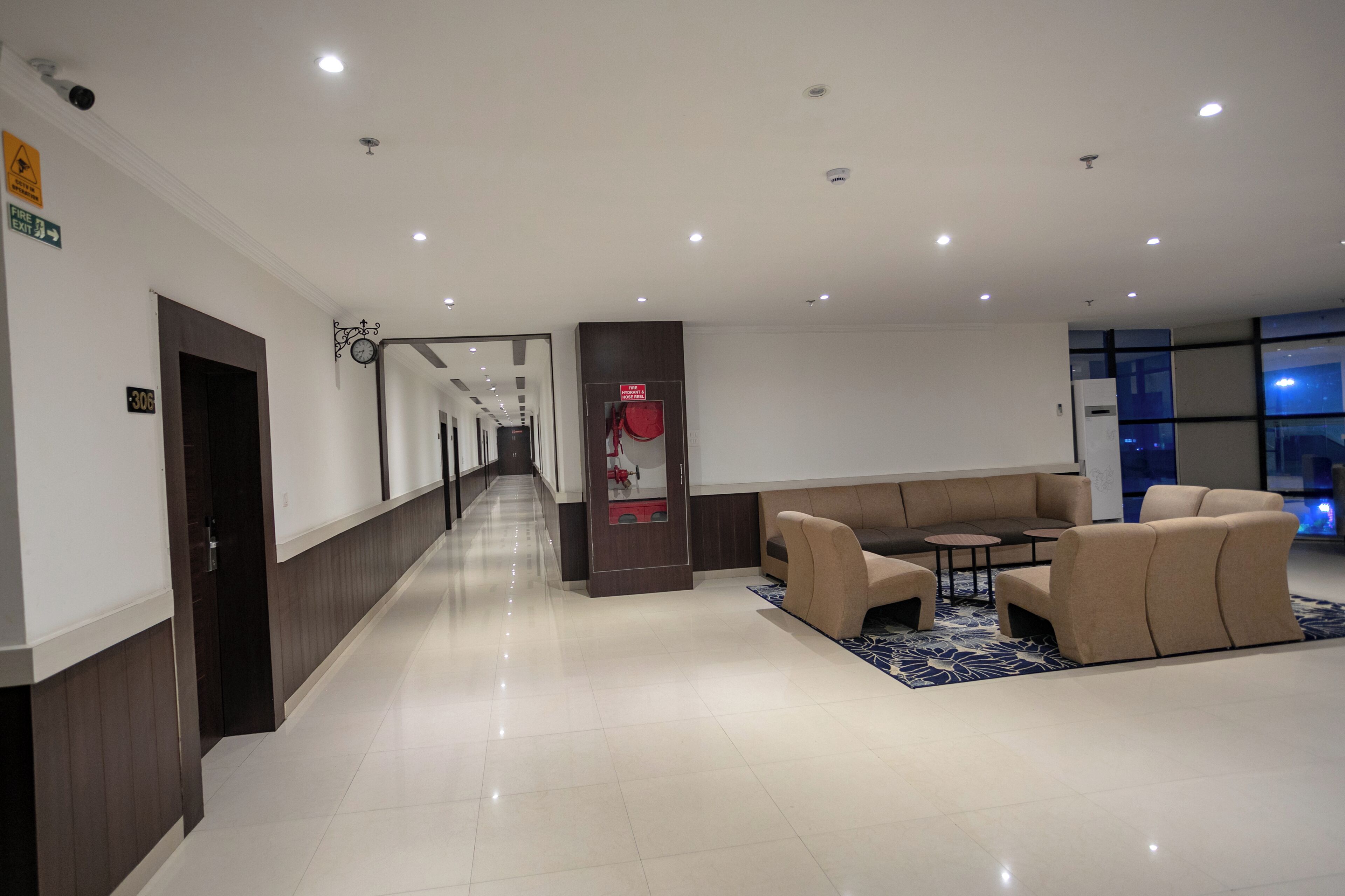 Um saguão de hotel moderno com um longo corredor, uma mesa de recepção, uma área de estar com sofás e um sinal de saída de incêndio.
