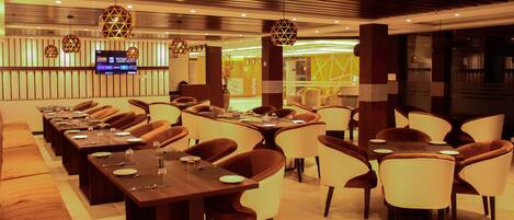 A modern dining area with long tables and chairs, overhead lighting, and a television screen.