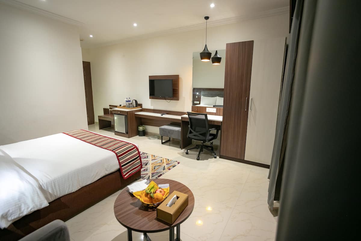 Quarto de hotel com cama, escrivaninha, cadeira, televisão e uma pequena mesa com caixa de lenços e frutas.