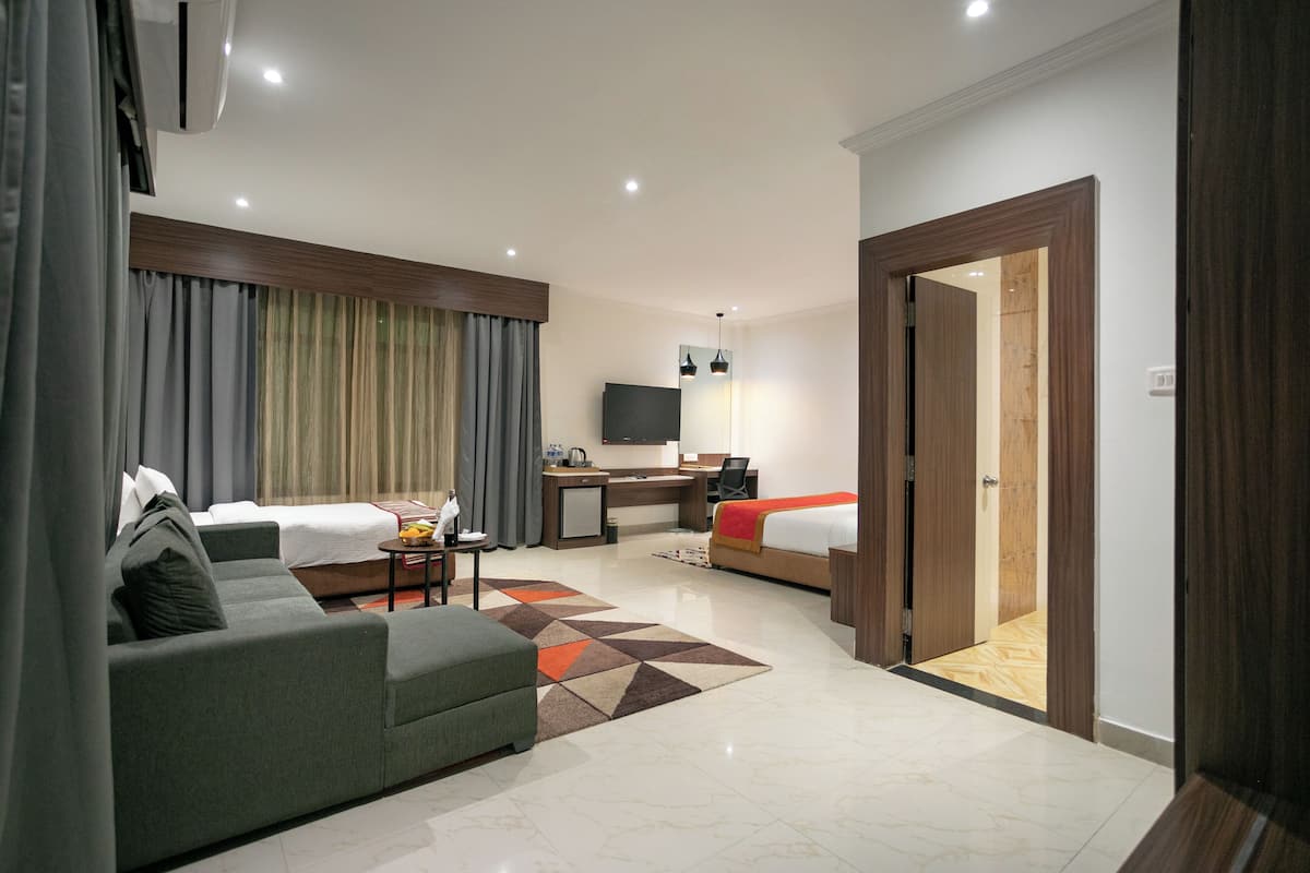 Quarto de hotel moderno com sofá, duas camas individuais, uma escrivaninha com cadeira, televisão e porta de madeira.