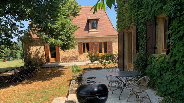 Une terrasse en pierre avec un barbecue, des sièges d’extérieur et un bâtiment en pierre doté de volets en bois.