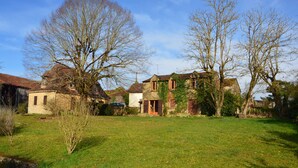 Une maison traditionnelle en pierre, dotée d’une pelouse bien entretenue, entourée d’arbres majestueux et baignée par un ciel dégagé.