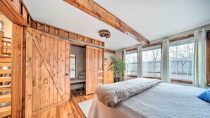 A bedroom with a wooden floor, a bed with a quilt, a large window with curtains, and a wooden wardrobe.