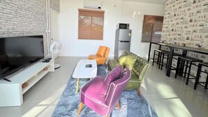 A modern living room with a flat-screen TV, a sofa, a chair, and a dining area with a table and chairs. - Homerent @ Sariam Lake Chiang Mai (San Pa Thong)