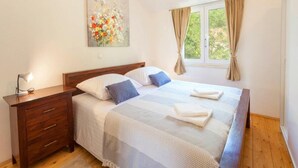 Een slaapkamer met een bed, een nachtkastje, een lamp, een schilderij en een raam met gordijnen.