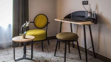 Habitación de hotel pequeña y funcional, con una mesita redonda, un sillón amarillo, un taburete y un escritorio con silla fijado a la pared.