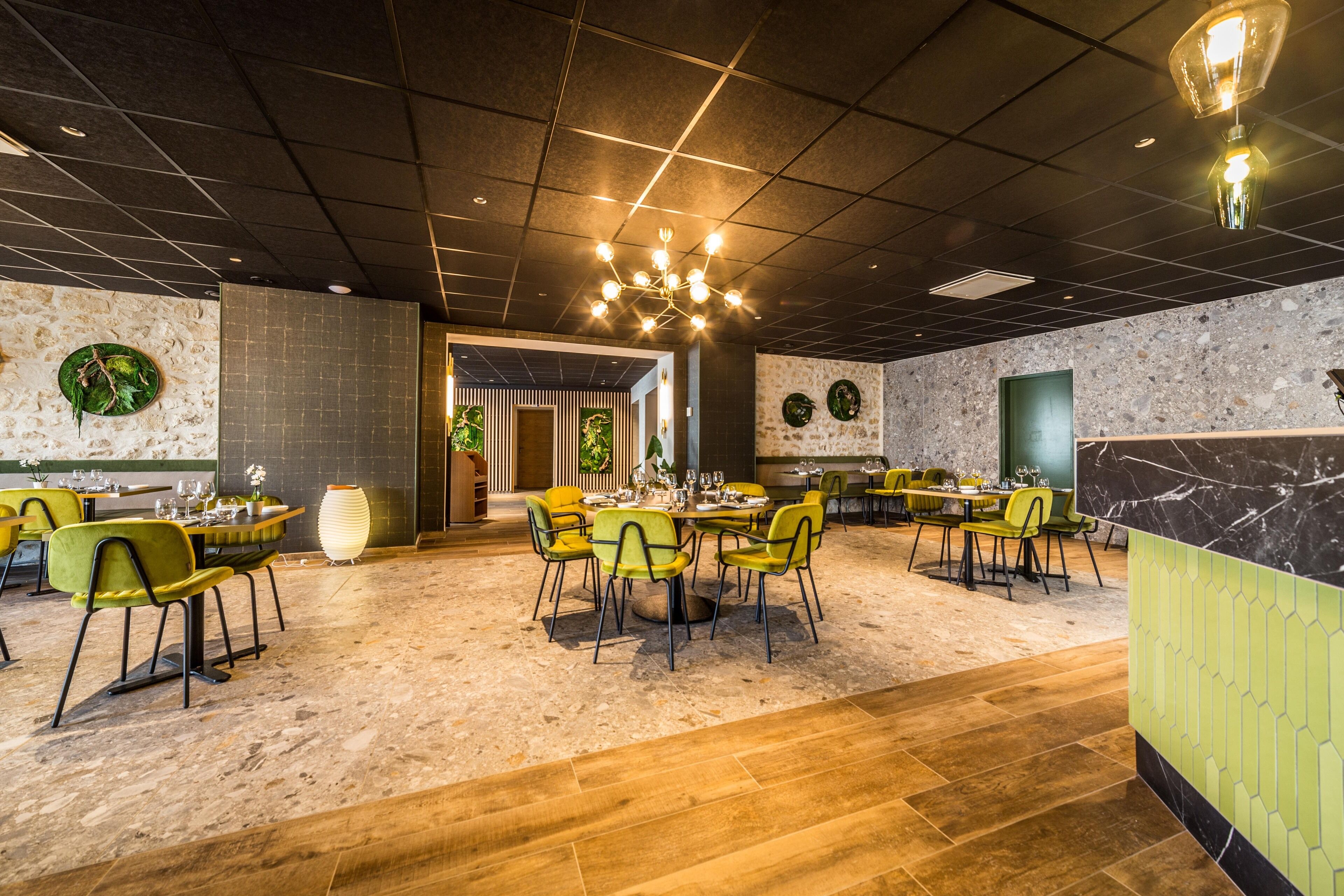 A spacious restaurant with multiple dining areas, each featuring round tables and green chairs. The interior includes a mix of stone and wood flooring, and decorative wall elements.
