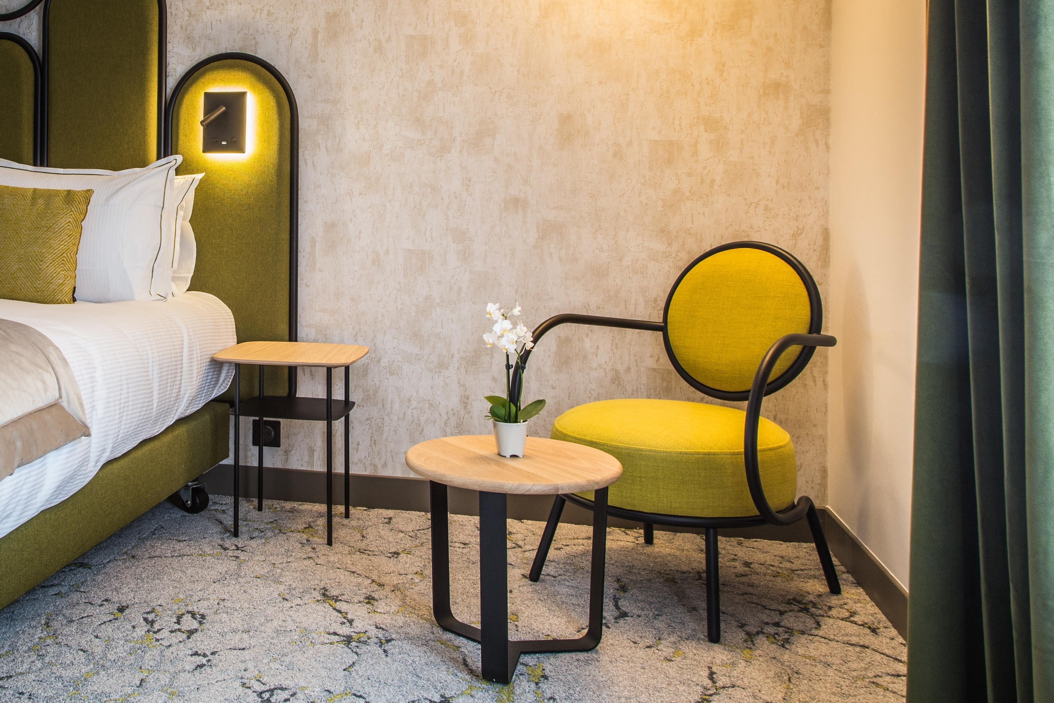 A hotel room with a bed, a small round table with a plant, a yellow chair, and a nightstand.