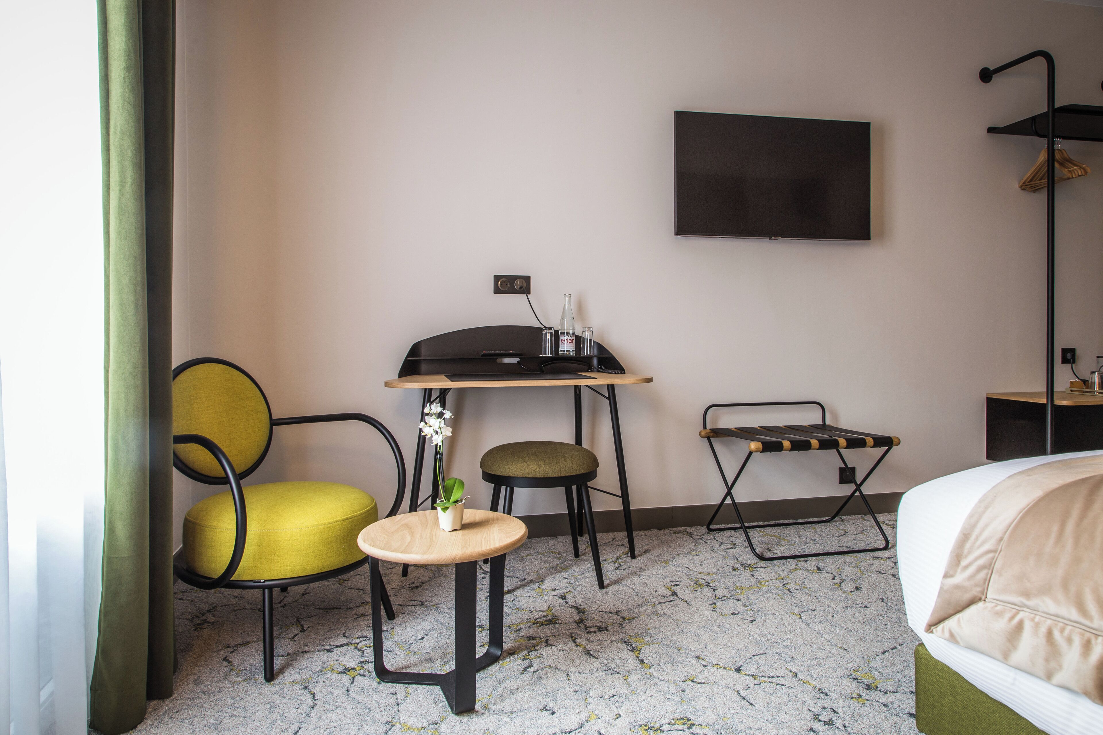 Una habitación de hotel con cama, un escritorio y una silla, una mesita con una planta, un televisor montado en la pared y una lámpara de pie.