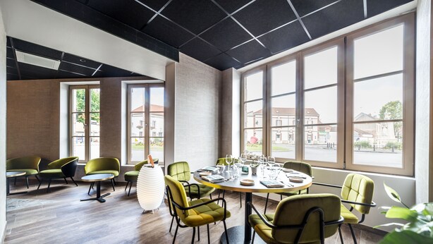 A modern dining area with a round table set for six, surrounded by green upholstered chairs. Large windows provide a view of the outside.