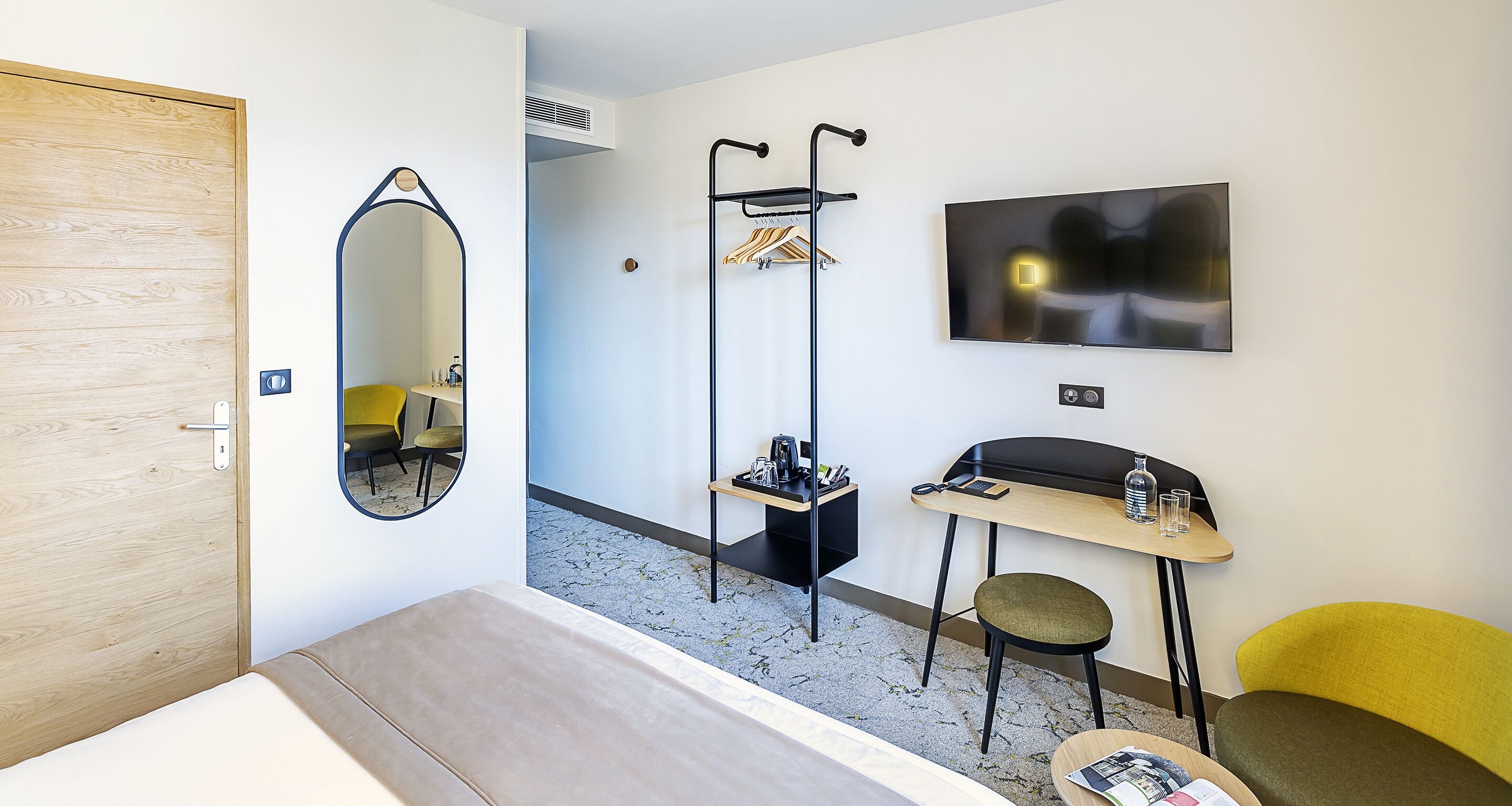 A hotel room with a wooden door, a mirror, a coat rack, a TV, a small table with a chair, and a bed.