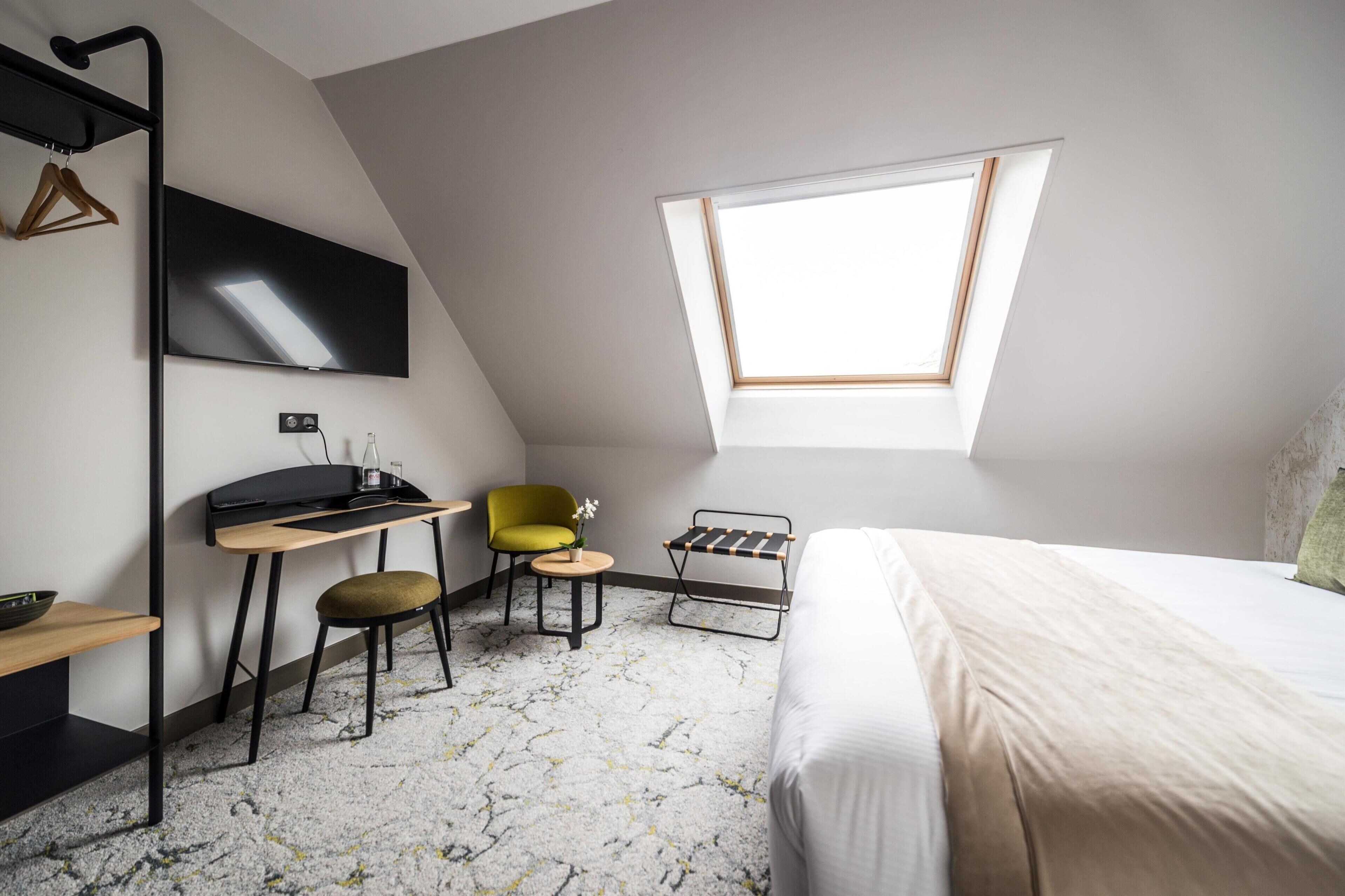 A modern bedroom with a skylight, a bed, a desk, a chair, and a small table with a plant.