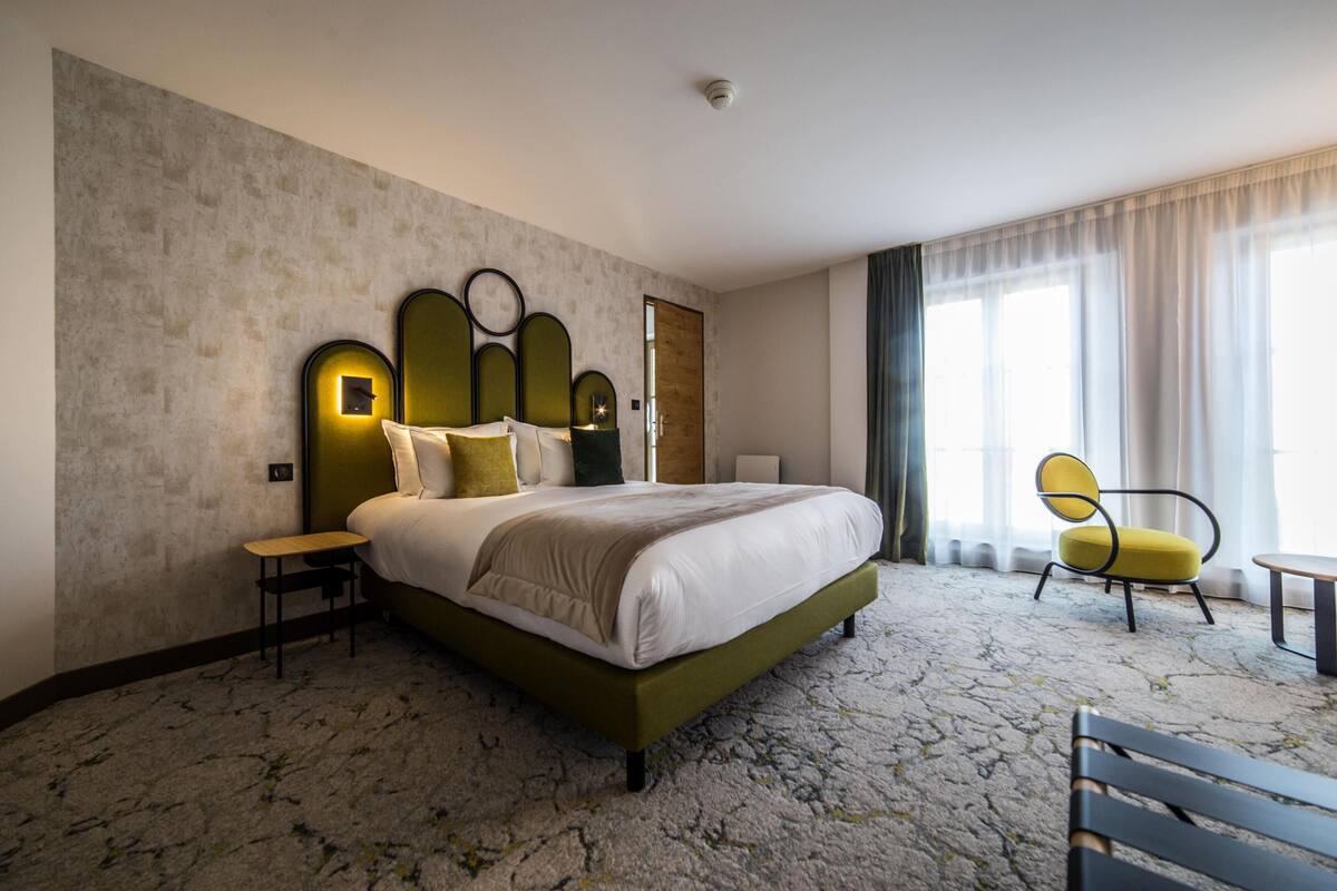 Ein Hotelzimmer mit einem großen Bett, einem Nachttisch, einem Sessel und einem Fenster mit Vorhängen.