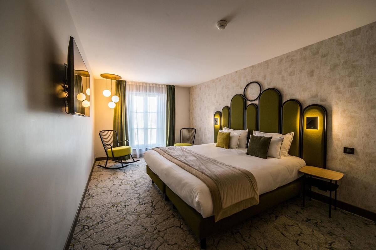 Ein Hotelzimmer mit einem großen Bett, einem Schreibtisch, einem Sessel, einem Fernseher und einem Fenster mit Vorhängen.