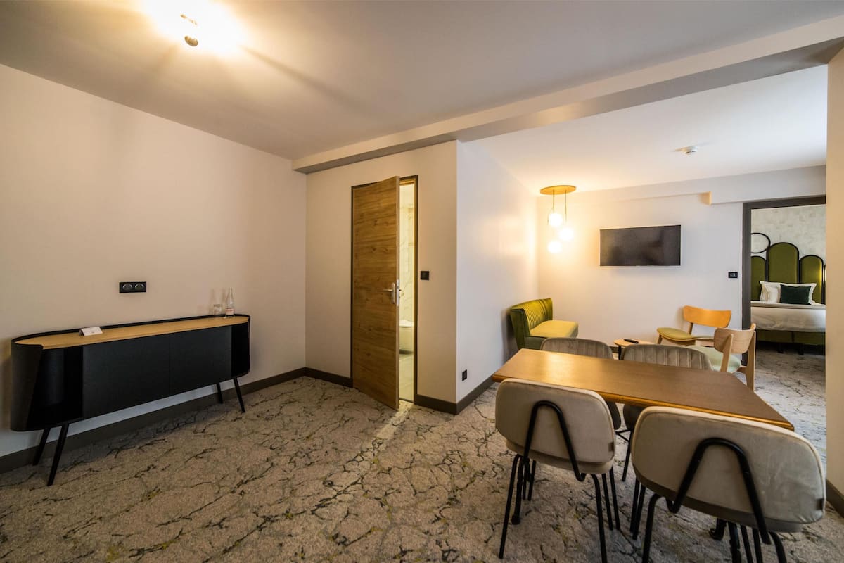 Ein Hotelzimmer mit Holztür, einem schwarzen Konsolentisch, einem Esstisch mit vier Stühlen, einer kleinen Couch und einem an der Wand befestigten Fernseher.