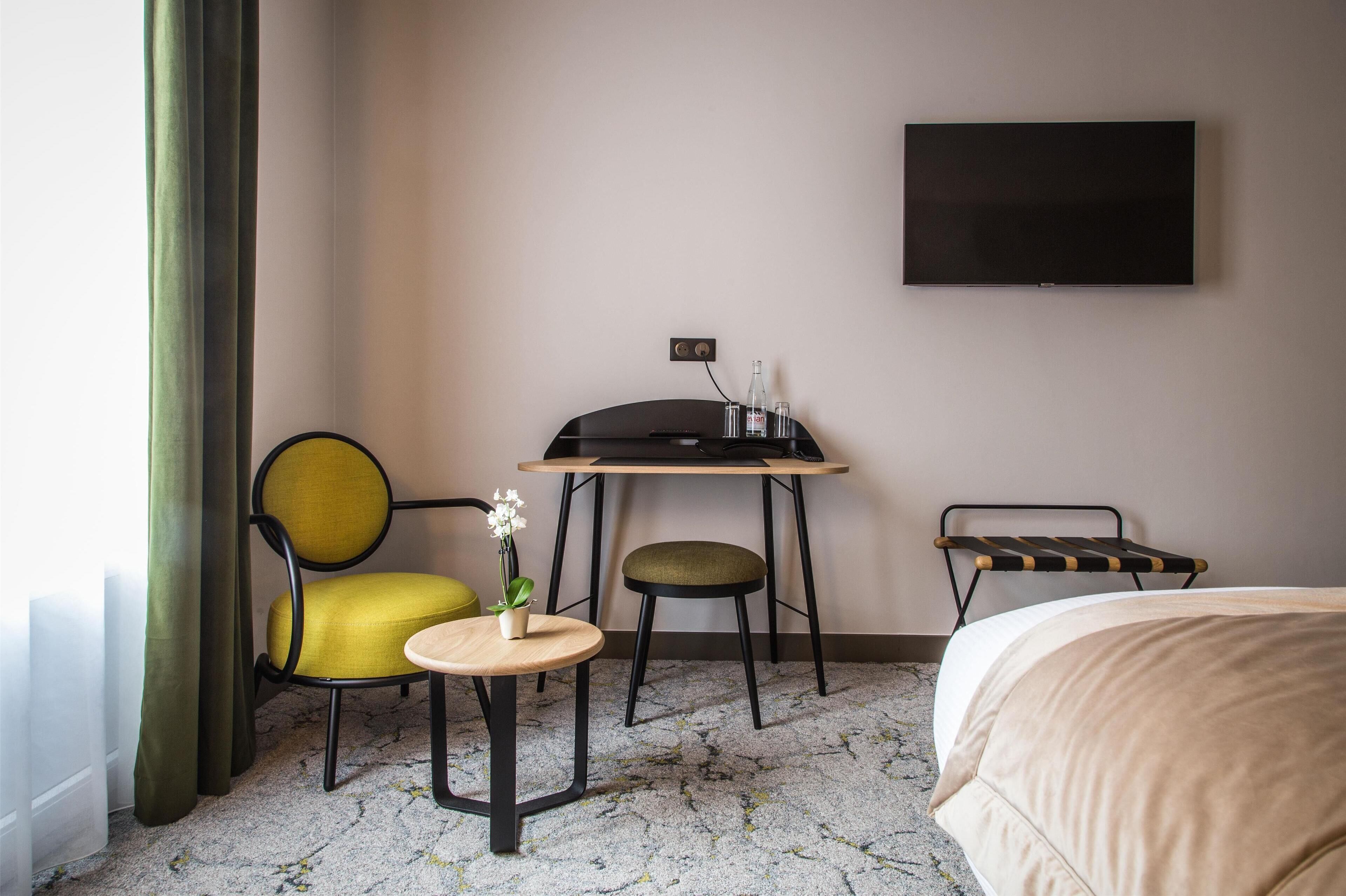 A hotel room with a bed, a desk with two chairs, a small table with a plant, and a TV on the wall.