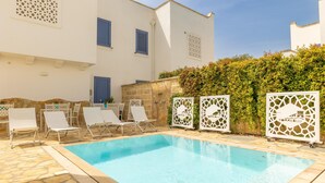 Una piscina con sillones de descanso y paneles decorativos, rodeada de un muro de piedra y follaje.