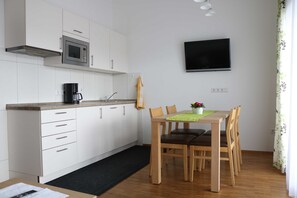 A modern kitchen with white cabinets, a microwave, and a coffee maker. There is a dining table with chairs and a TV mounted on the wall.