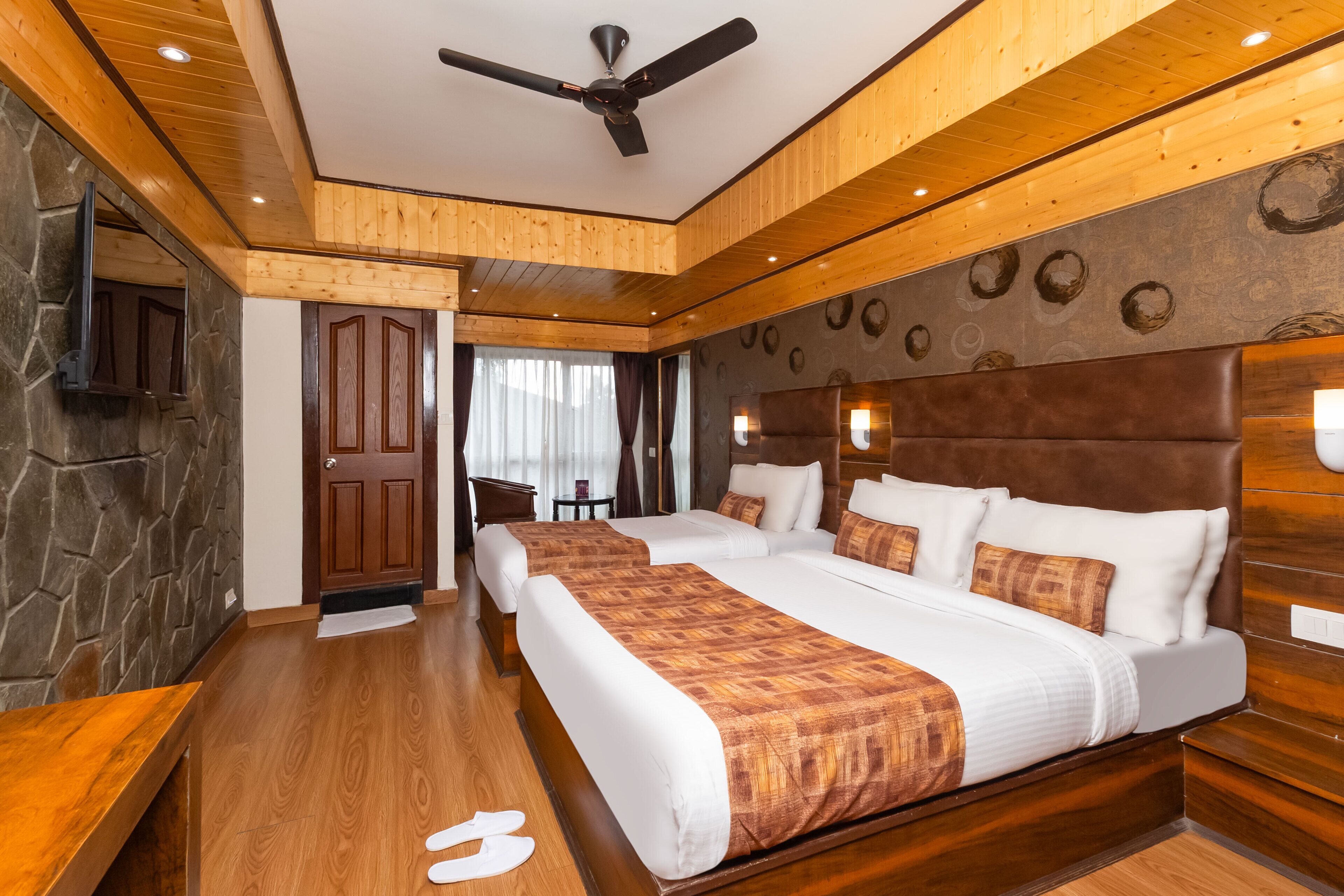 Ein Hotelzimmer mit zwei Betten, Holzboden, einer Wand aus Stein und einem Deckenventilator.