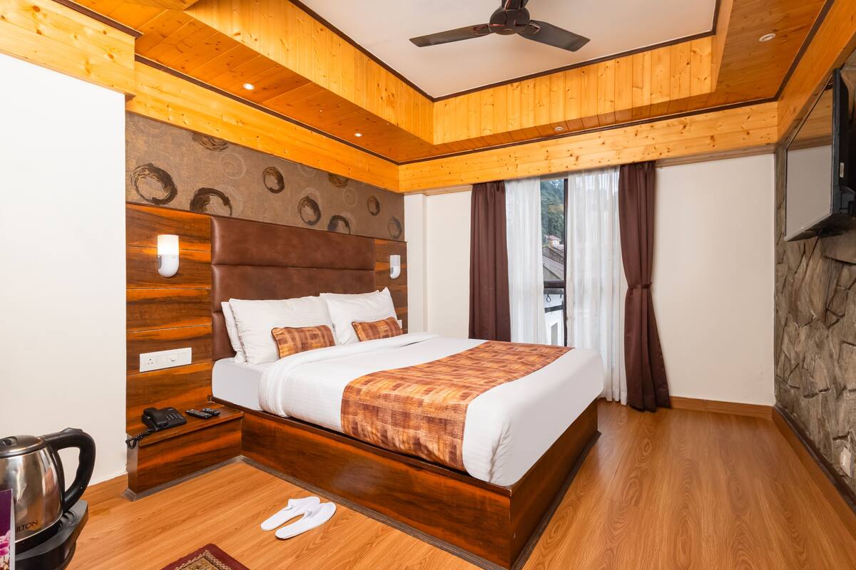 A modern hotel room with a wooden ceiling, a large bed with a headboard, a nightstand, a kettle, and a view of the outdoors through the window.
