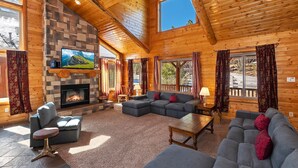 A log cabin interior with a stone fireplace, a large gray sectional sofa, and a wooden dining table.