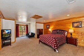 A bedroom with a wooden wall, a bed with a patterned bedspread, a television on a stand, and a window with curtains. - Serenity Now Lodge by Destination Big Bear: Luxury Log Home in Castle Glen Estates! Hot Tub! Pool Table! Ping Pong! (Big Bear Lake)