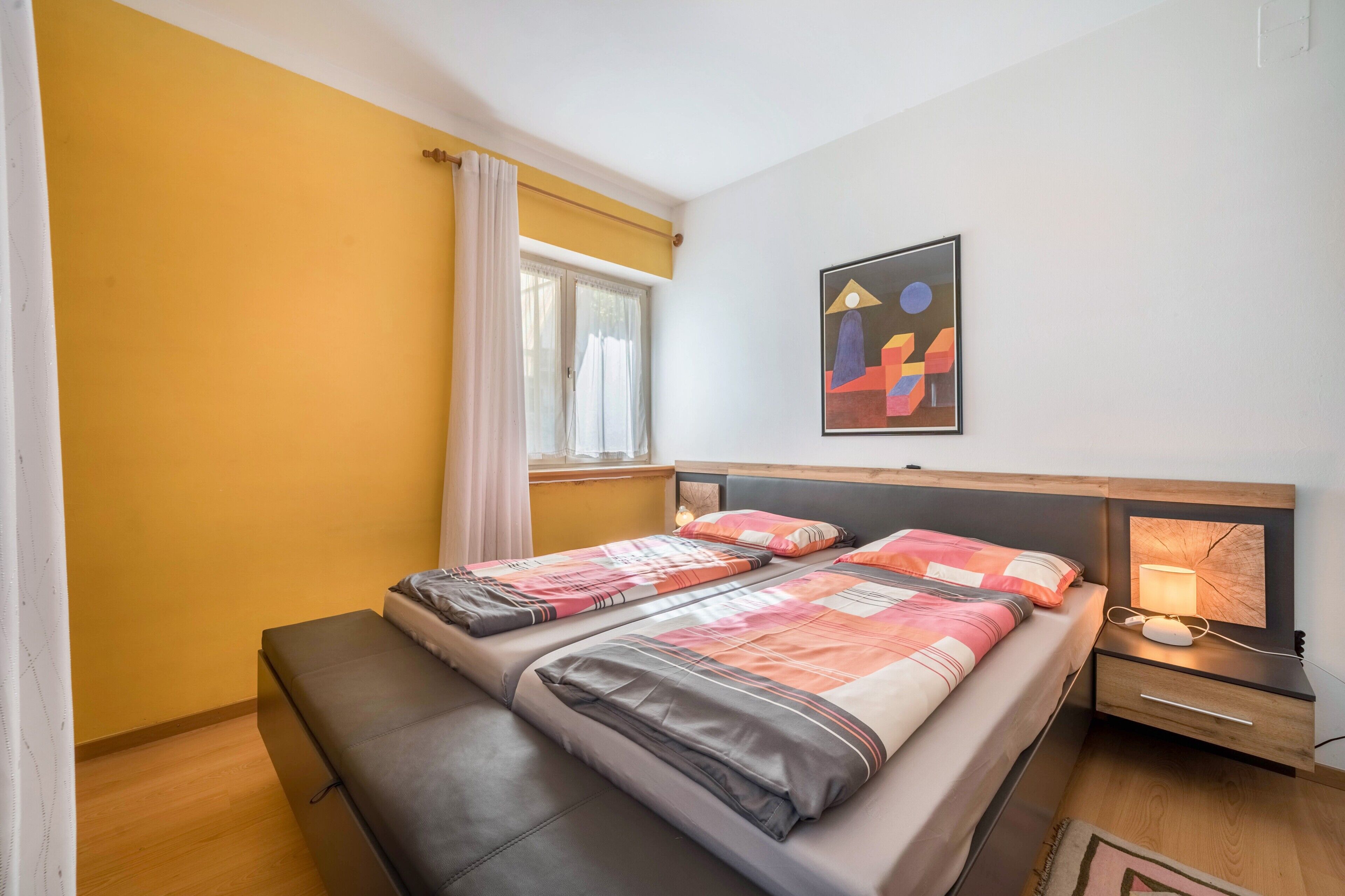 Een kamer met een bed, een bankje, nachtkastje, lamp en een schilderij aan de muur.