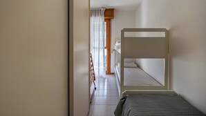Una habitación pequeña con una cama individual superpuesta, una puerta de madera y una ventana con cortinas translúcidas.