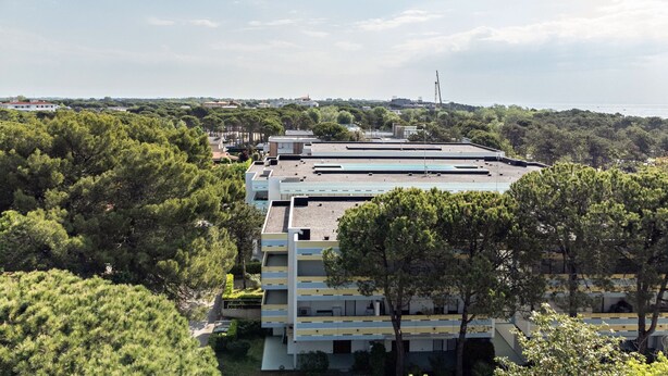 Un edificio de varios pisos con techo plano, rodeado de abundante vegetación y un cielo despejado.