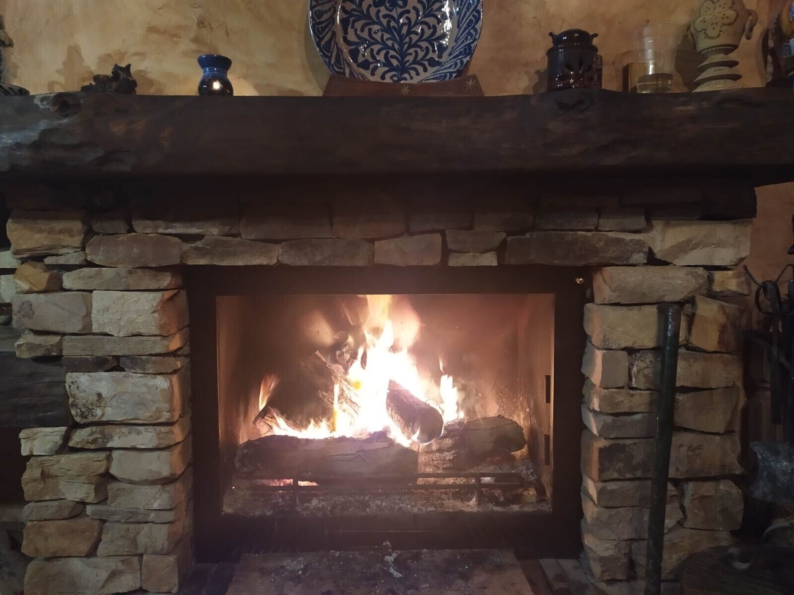 Una chimenea de piedra con fuego encendido, un jarrón azul decorativo sobre la repisa y un estante de madera rústica arriba.