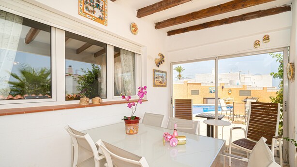 Un coin repas avec une table en verre, des chaises blanches, une plante en pot et une vue sur les bâtiments et la piscine à l’extérieur.