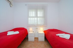 Een kamer met twee bedden, elk voorzien van een rode sprei en een witte handdoek, twee nachtkastjes met lampen, een raam met witte gordijnen en een airconditioner aan de muur.