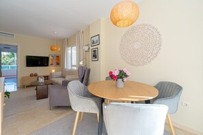 Una sala de estar moderna con un sofá, dos sillas grises, una mesa redonda de madera con un florero y un televisor de pantalla plana, además de un elemento decorativo en la pared.