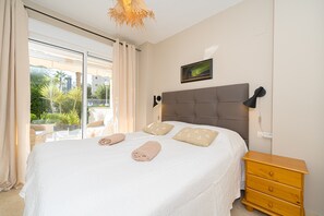Een slaapkamer met een bed, een nachtkastje, een raam met gordijnen en uitzicht op groen buiten.