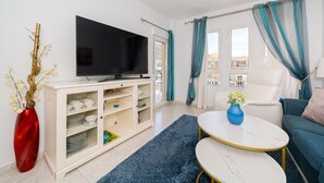 Een moderne woonkamer met een flatscreen televisie, een witte kast met planken, een blauw vloerkleed, een witte bank en een blauwe vaas.