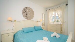 Een slaapkamer met een bed, nachtkastje, dressoir, raam met gordijnen en een kunstwerk aan de muur.