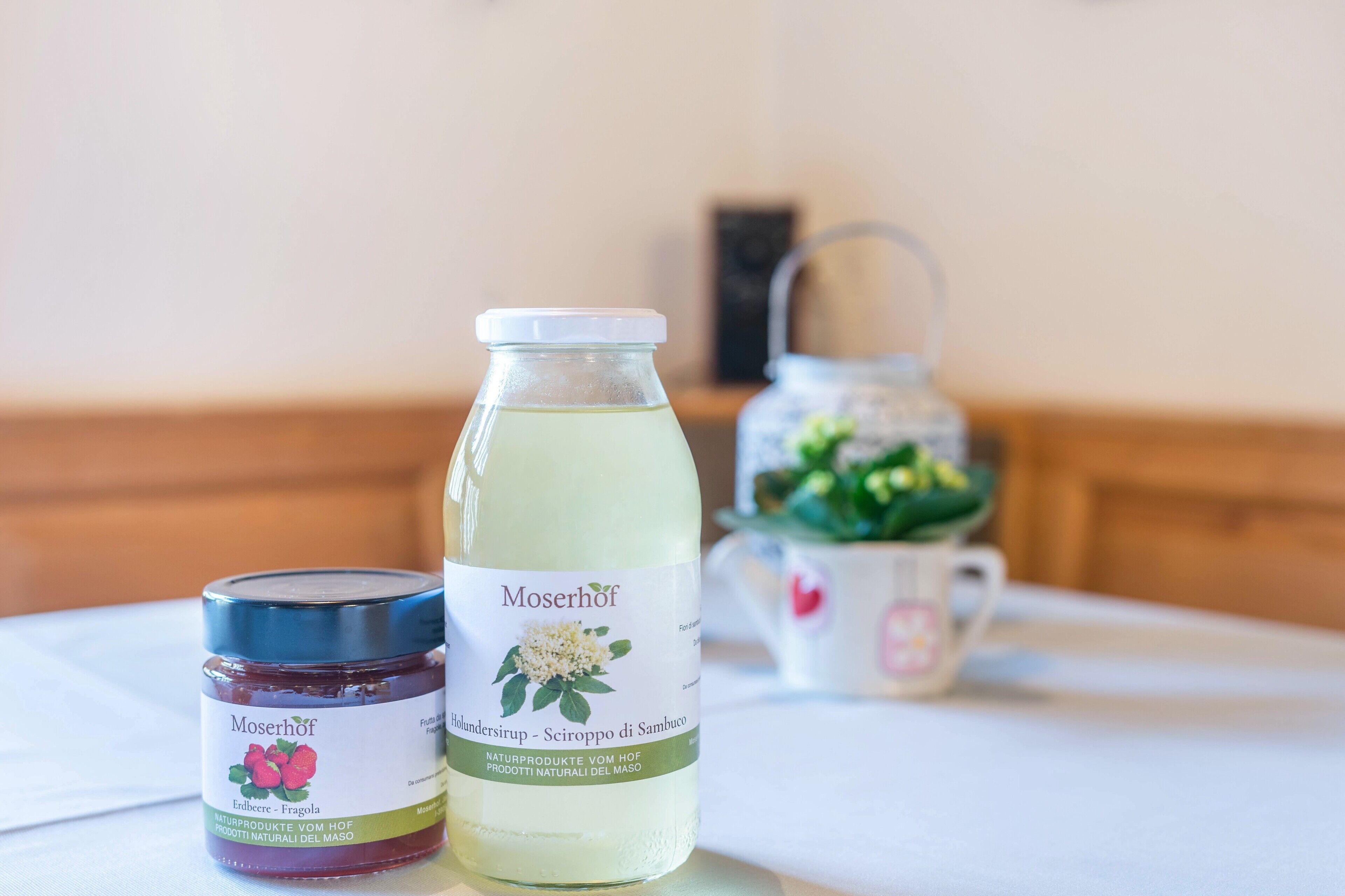 Ein Tisch mit einer Flasche Moserhof-Kräutersirup, einem Glas Moserhof-Beerenkonfitüre und einem Teekessel mit einer Pflanze.